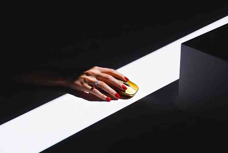 Eine weibliche Hand mit leuchtend rotem Nagellack und einem silbernen Ring auf dem Ringfinger ruht auf einer kleinen, goldenen Computermaus.